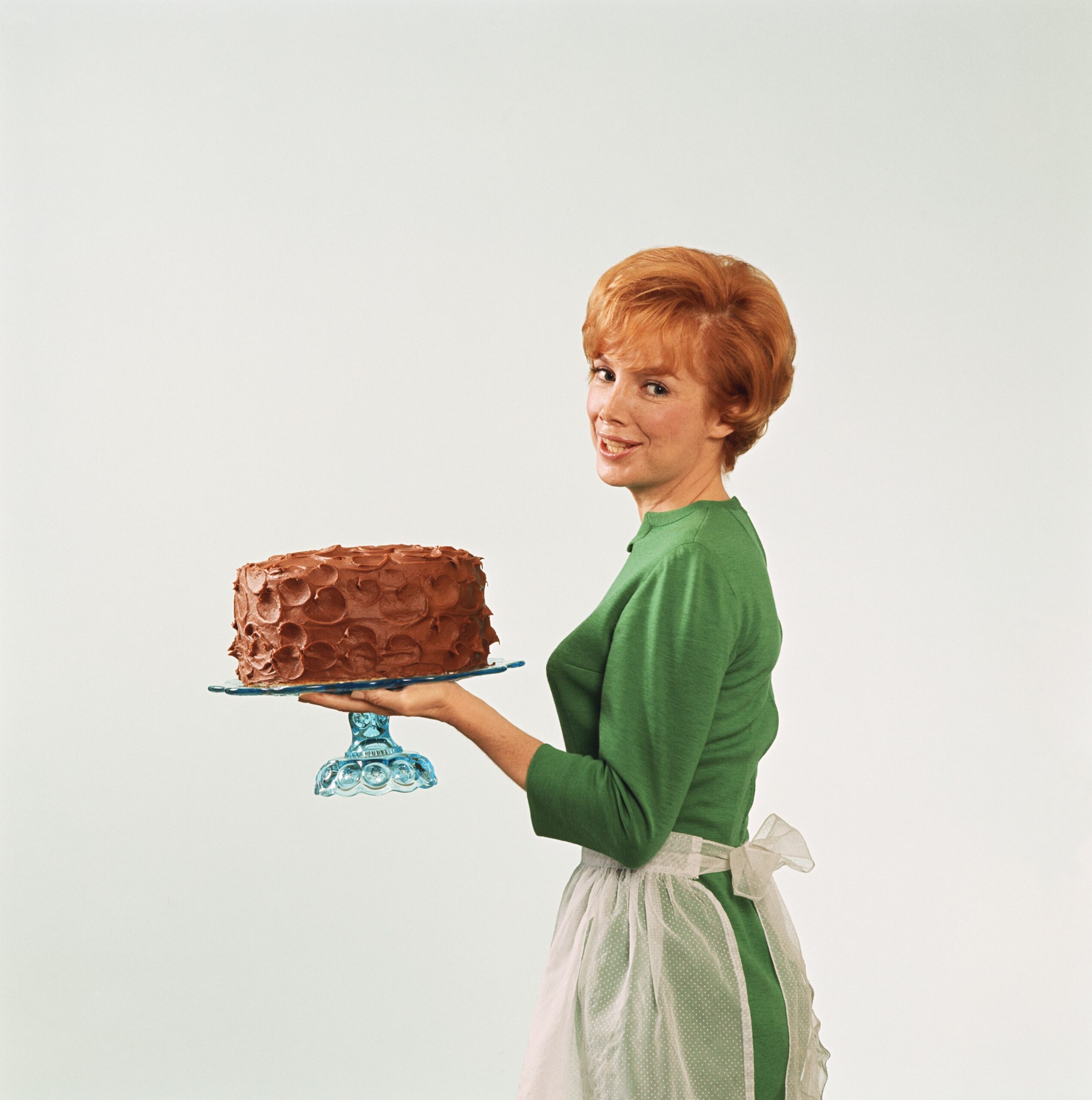 Woman in a green dress and apron holding a chocolate cake.